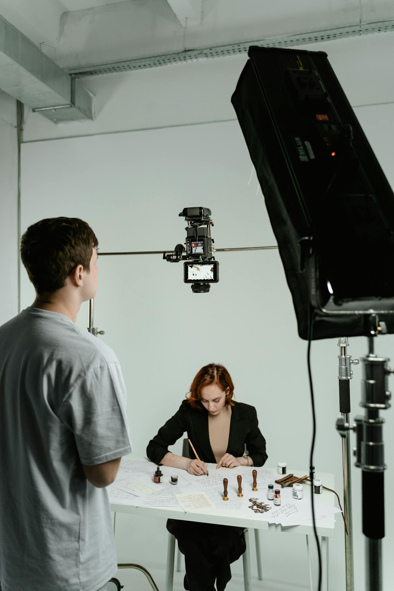 A printed contact sheet of 12 medium-format frames laid out on a working table next to a calibrated monitor, the contact sheet marked with grease pencil.